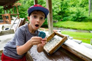 an excited kid finds a blue gemstone while sifting through earth and sand with a screen at Gem Mountain